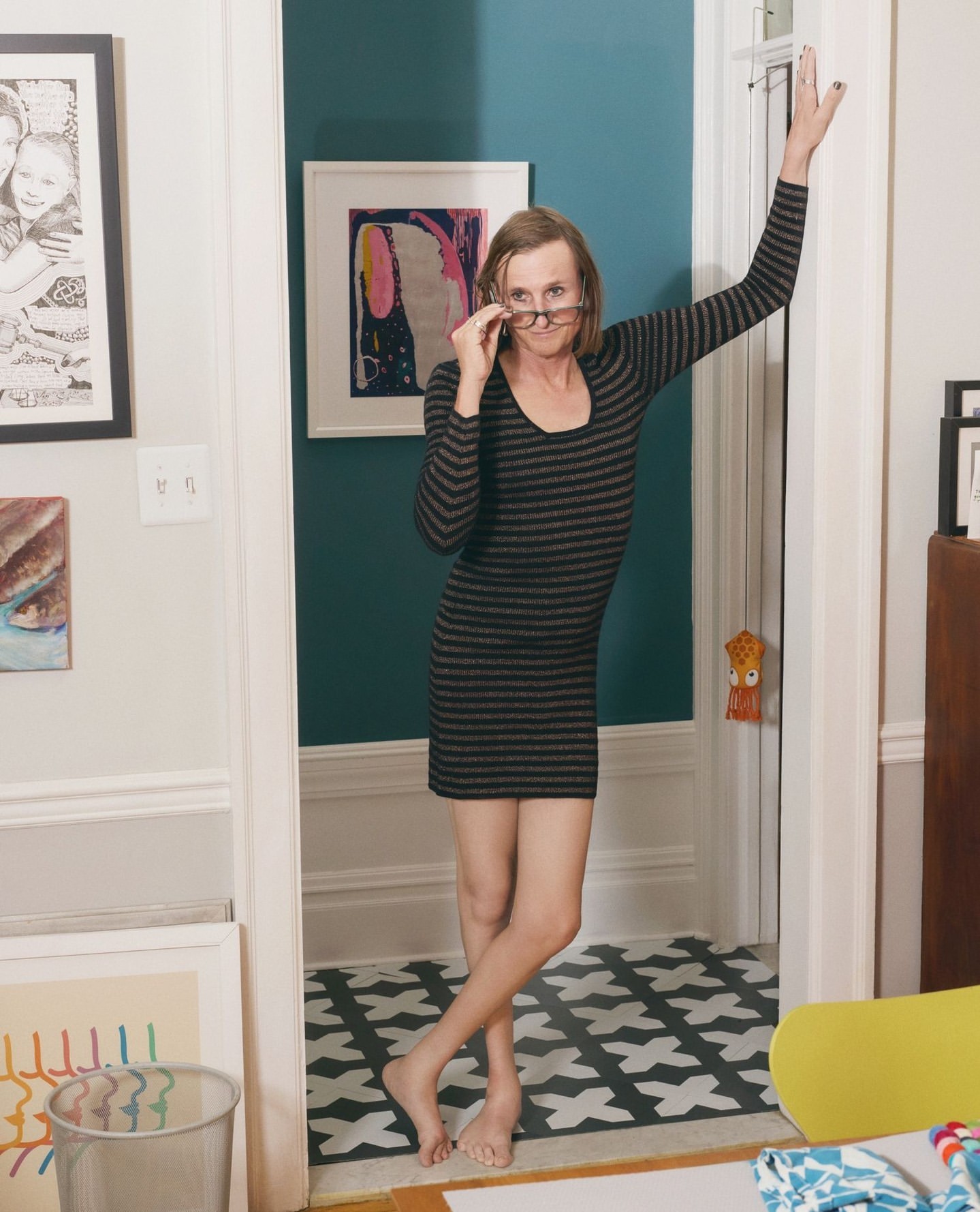 Woman in striped dress poses in doorway with eyeglasses, framed art on teal walls, and geometric floor pattern in residential setting.
