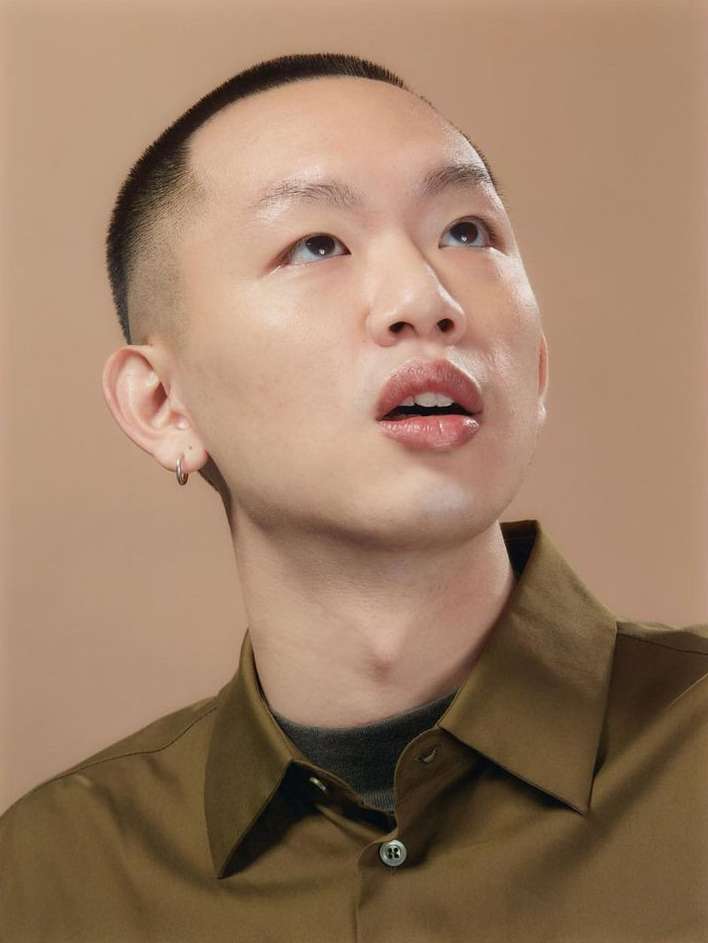 Portrait of a young Asian man with short dark hair wearing a green button-down shirt, looking up with an open mouth against a beige background.