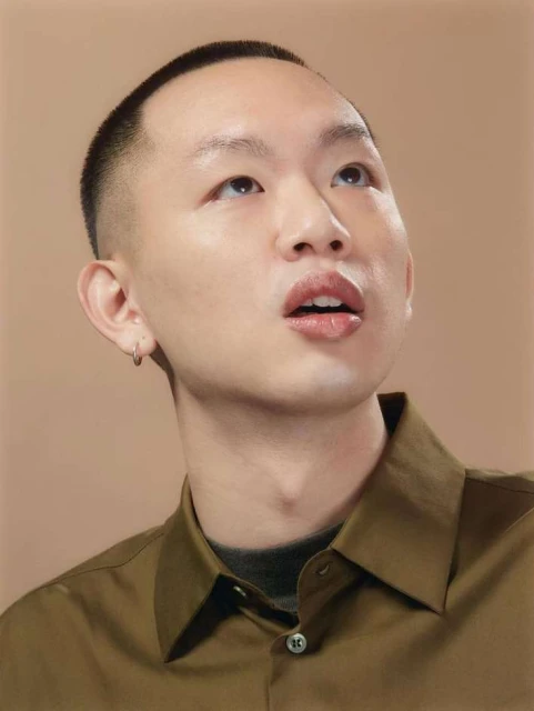 Portrait of a young Asian man with short dark hair wearing a green button-down shirt, looking up with an open mouth against a beige background.