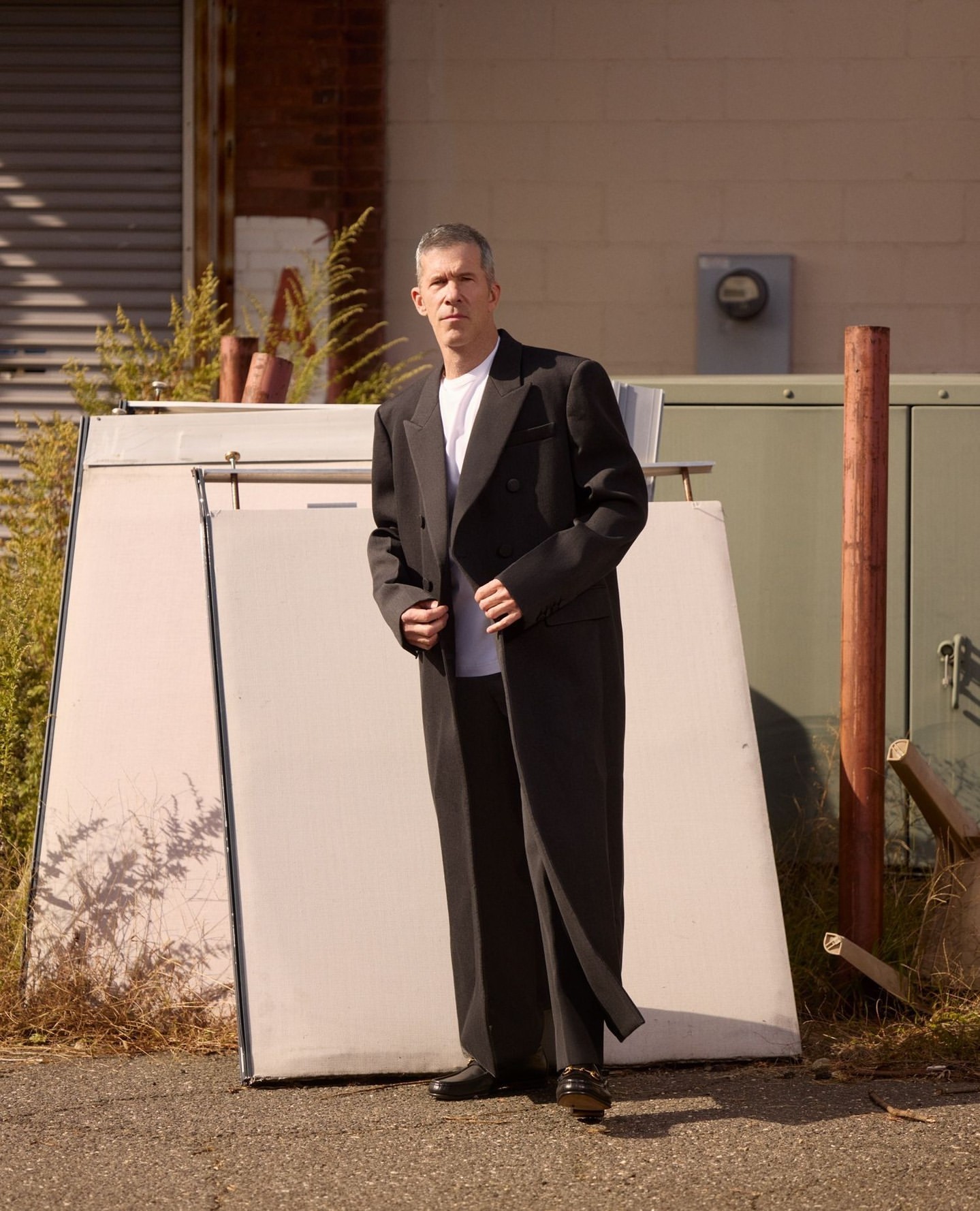A man in a black suit stands in an urban setting near a building, leaning against a large white panel with an expressionless face.