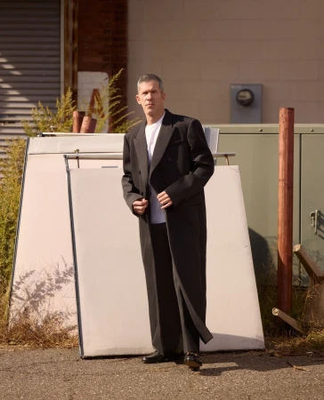 A man in a black suit stands in an urban setting near a building, leaning against a large white panel with an expressionless face.
