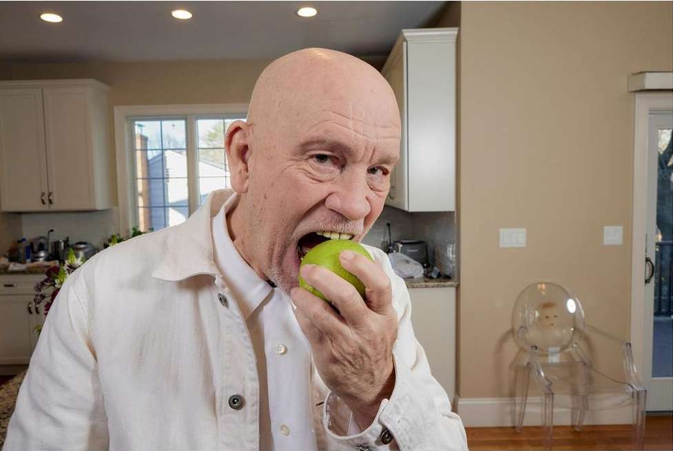 Close-up of an adult male with a bald head indoors, eating a green apple while wearing a white jacket in a kitchen.