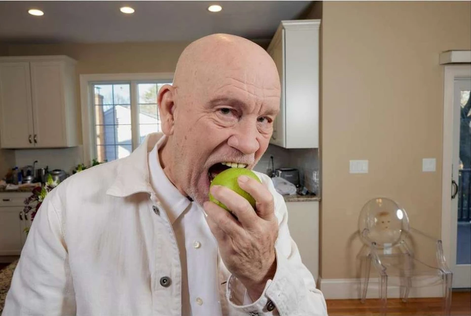 Close-up of an adult male with a bald head indoors, eating a green apple while wearing a white jacket in a kitchen.