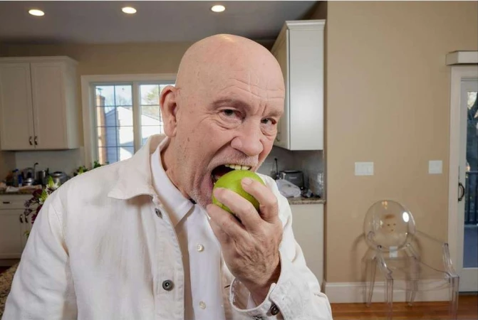 Close-up of an adult male with a bald head indoors, eating a green apple while wearing a white jacket in a kitchen.