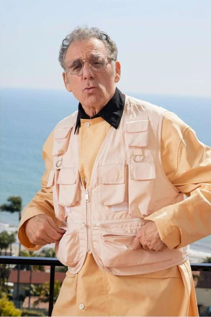 Elderly man in glasses, wearing a fishing vest over a peach shirt, stands outdoors with a sea view in the background on a sunny day.