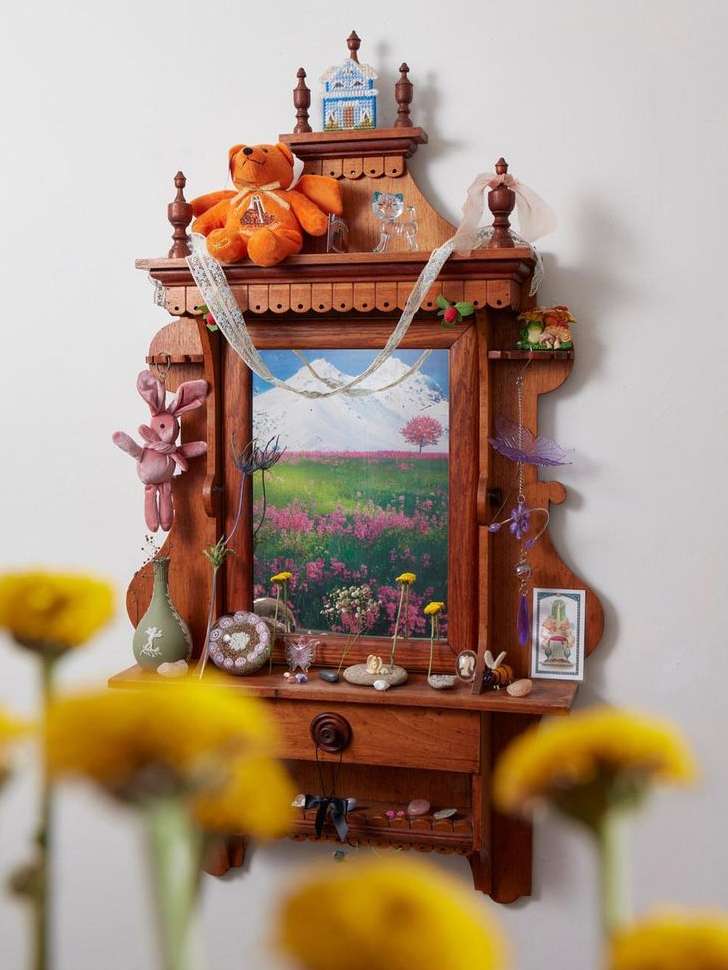 Wooden display shelf with a landscape painting, a teddy bear, and other small objects against a white wall, flowers in the foreground.