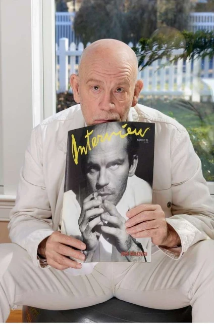 Portrait of a balding man holding an Interview magazine cover featuring a black-and-white photo of himself, indoors by a window.