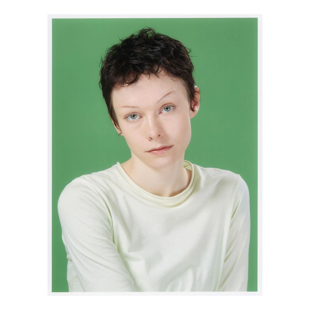 Portrait of a woman with short dark hair and fair skin, wearing a long-sleeved light green shirt, against a solid green background.