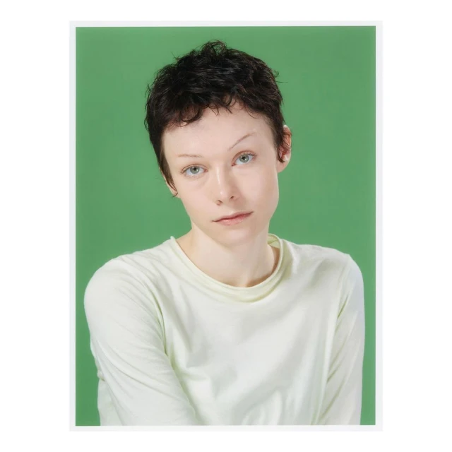 Portrait of a woman with short dark hair and fair skin, wearing a long-sleeved light green shirt, against a solid green background.
