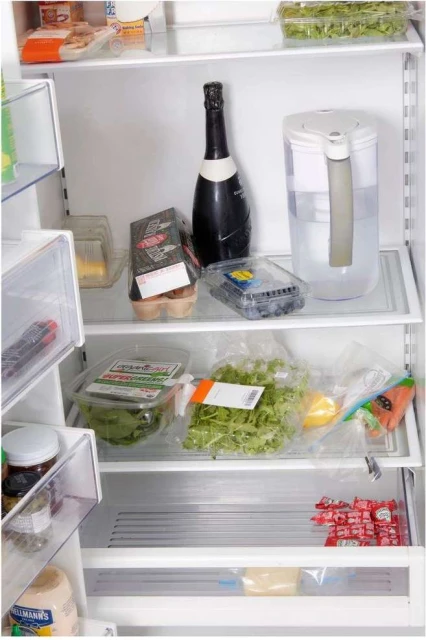 Inside of a refrigerator filled with food and drink items, including fruit, vegetables, eggs, sparkling wine, and a water filter pitcher.