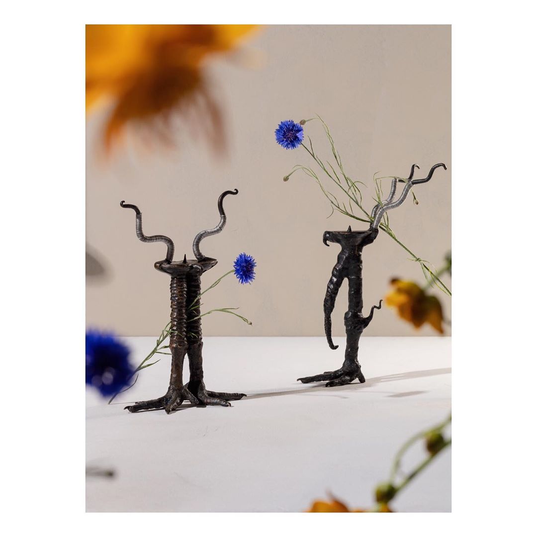 Two decorative pedestals hold single blue cornflowers against a neutral backdrop with soft, blurred yellow and gold floral elements.