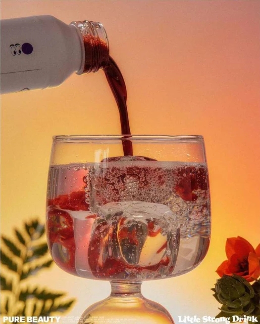 A glass of water with red liquid and ice being poured into it from a white bottle against a golden gradient background.