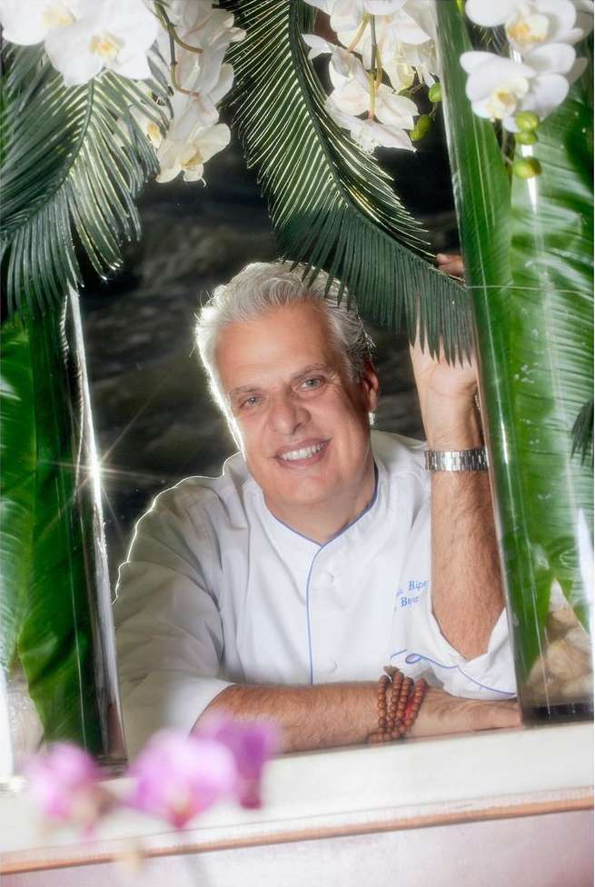 Smiling chef in a white coat surrounded by white orchids and green tropical leaves in an indoor setting with natural lighting.