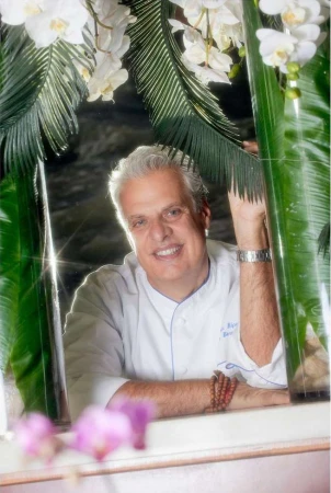Smiling chef in a white coat surrounded by white orchids and green tropical leaves in an indoor setting with natural lighting.