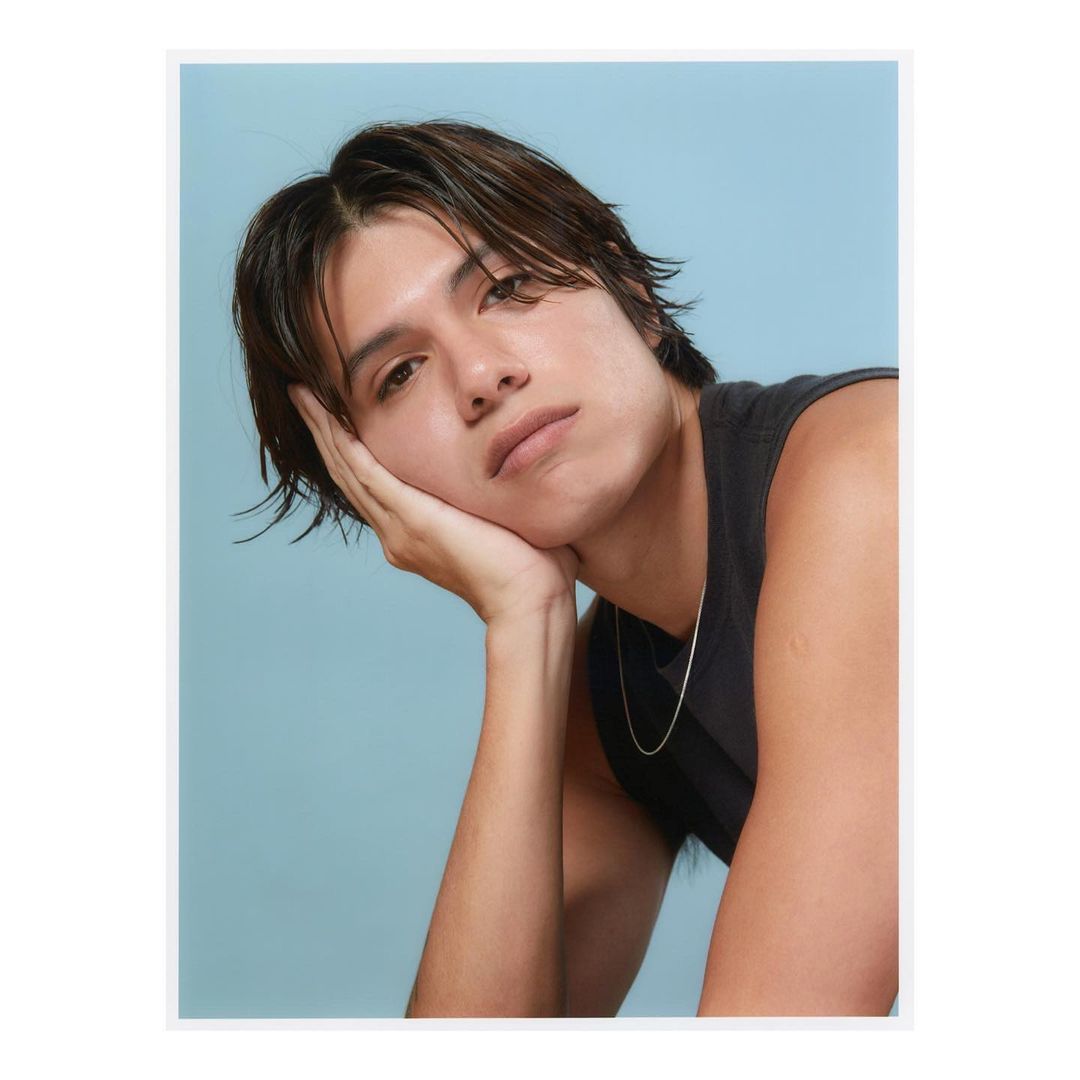 Portrait of a young adult male with dark hair resting his face on his hand, wearing a sleeveless top against a light blue backdrop.