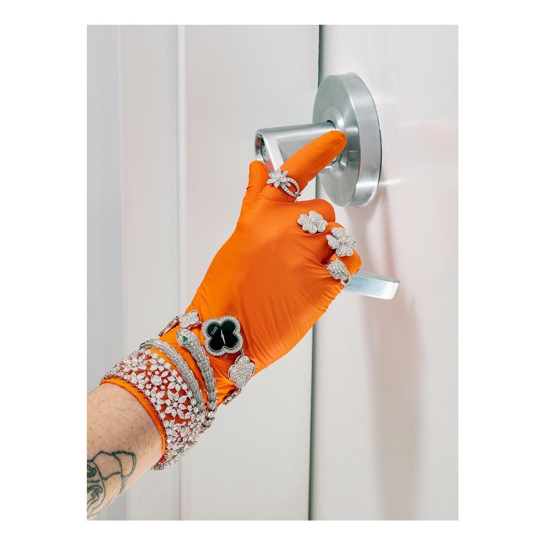 A hand wearing orange gloves and adorned with several diamond rings and bracelets pushes a door handle in a bright, indoor setting.