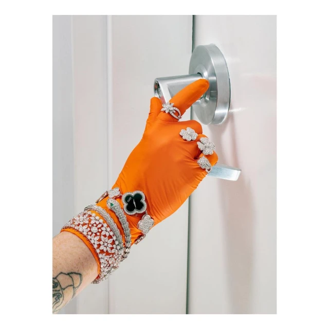 A hand wearing orange gloves and adorned with several diamond rings and bracelets pushes a door handle in a bright, indoor setting.