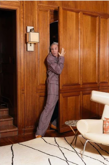 An adult male wearing a purple suit peers out from behind ajar wooden closet doors in a brightly lit wood-paneled room with modern furniture.