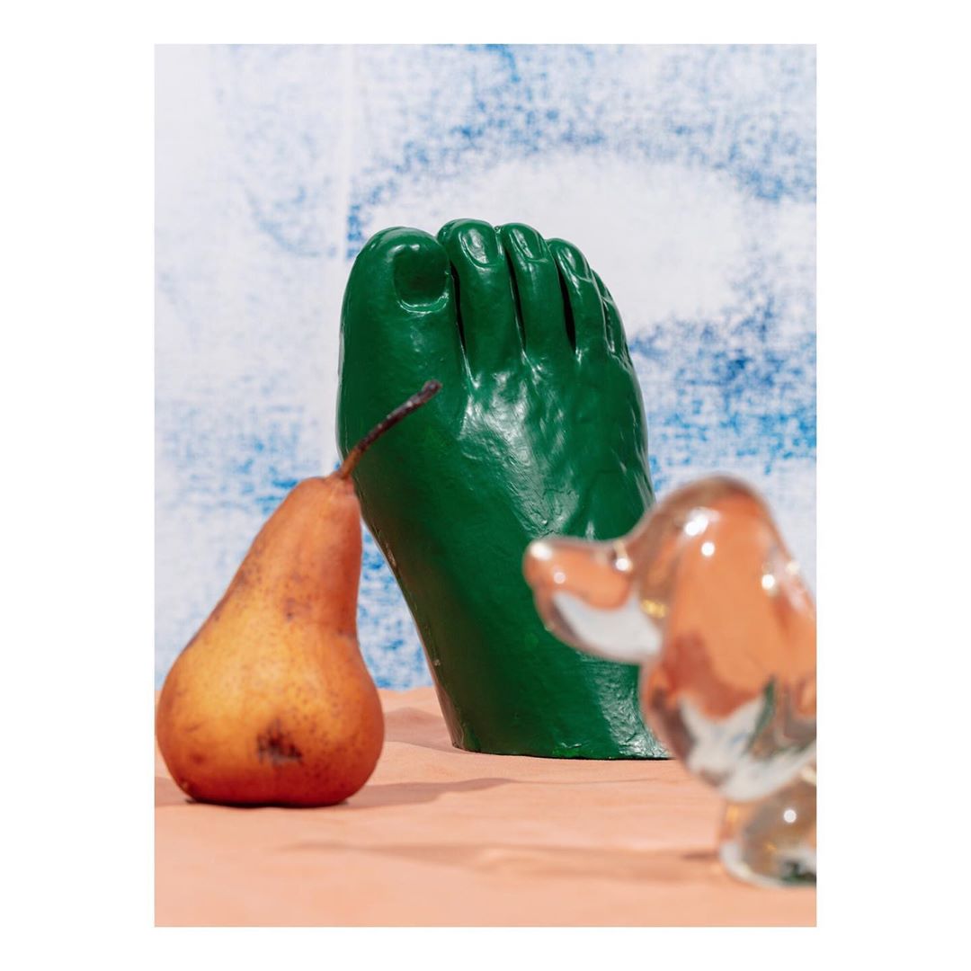 Surreal still life featuring a green plaster foot, a pear, and a glass animal figurine against a mottled blue and white background.