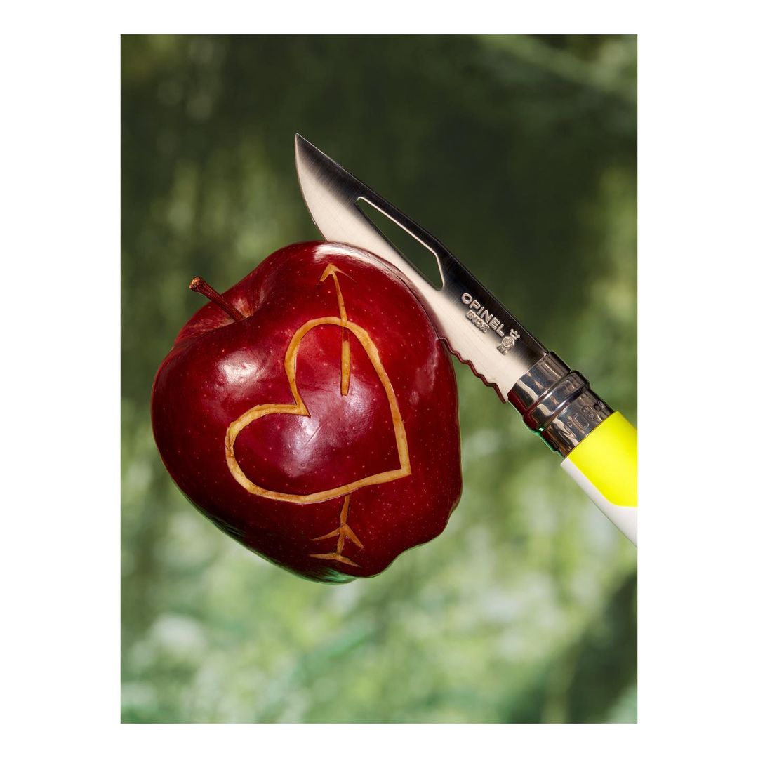 A red apple carved with a heart and arrow, alongside an Opinel knife with a yellow handle, set against a blurred green background.