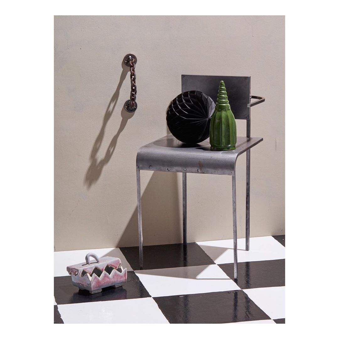 Still life photograph of a gray metal chair with a green glass bottle and black textured ball, plus geometric ceramic box, on a checkered floor.