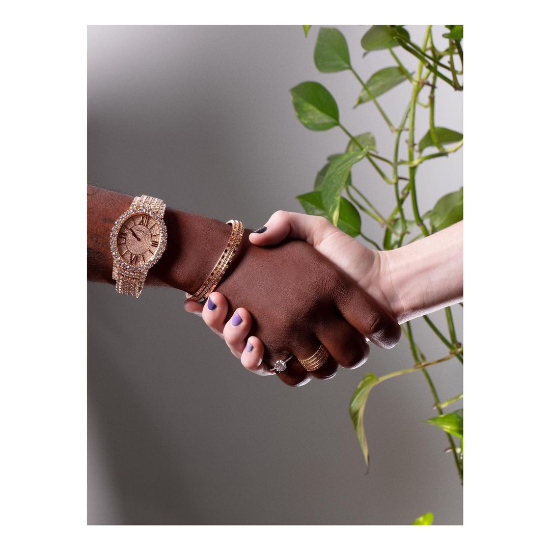 Close-up of two people of different ethnicities shaking hands, wearing rings and bracelets, against a grey background with plants.