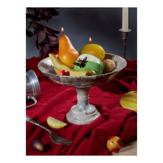 Still life photography of fruit bowl with candles on red fabric, including pear, lemon, banana, plum, cherry, walnut, and green dessert.