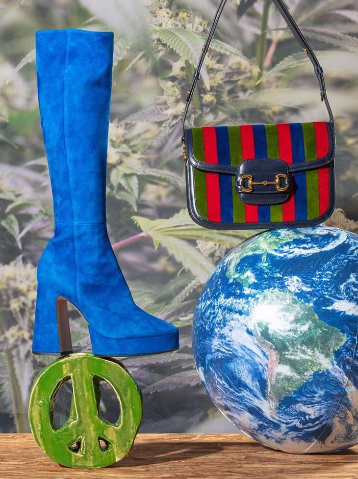Still-life studio shot of a blue platform boot, a Gucci striped bag, an earth globe, and a green peace-sign figure against a leaf background.