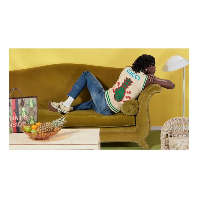 A man reclines on an olive-green couch in a fashion editorial. He wears jeans, a patterned vest, and platform shoes, with retro-inspired decor.