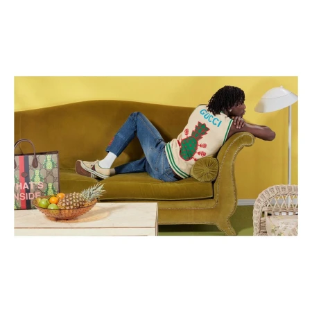 A man reclines on an olive-green couch in a fashion editorial. He wears jeans, a patterned vest, and platform shoes, with retro-inspired decor.