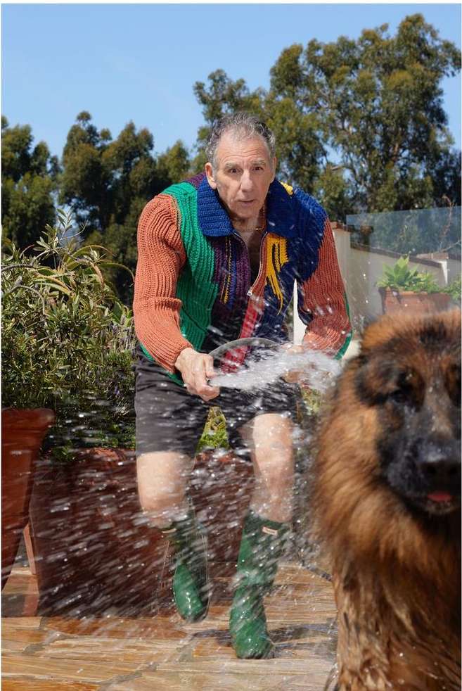Adult male in colorful sweater holding a hose, spraying water onto a large dog in an outdoor setting with foliage and blue sky.