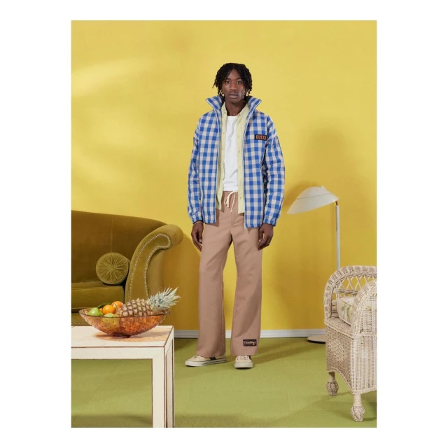 An adult man in a blue checkered jacket and beige pants stands indoors against a yellow wall with a sofa and lamp.