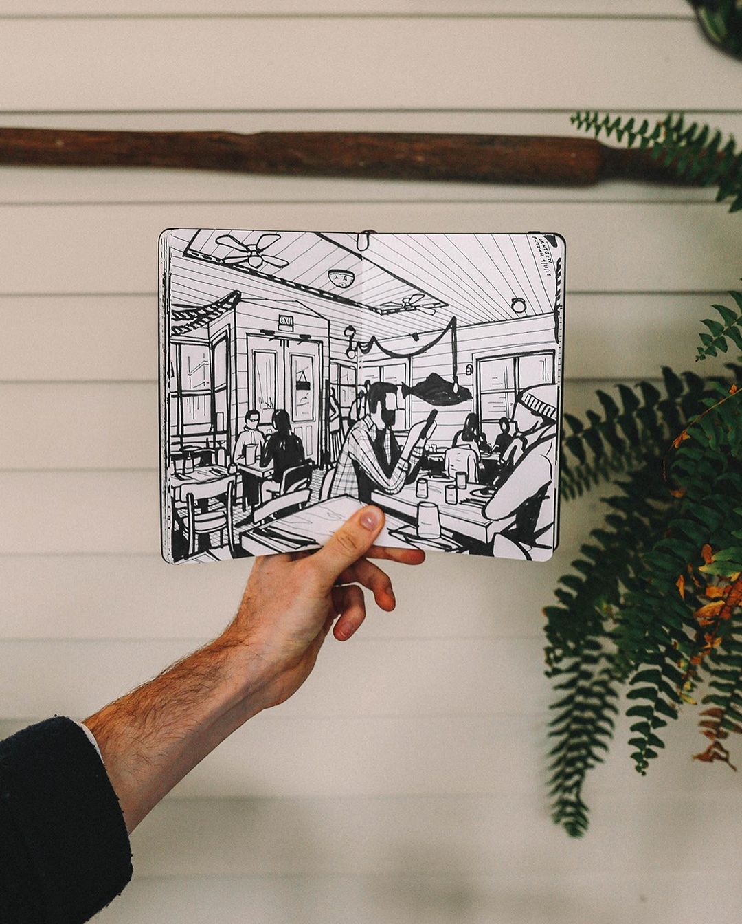 Hand holding a sketch of people in a restaurant, showing interior tables, chairs, and customers, set against a white wall and fern.
