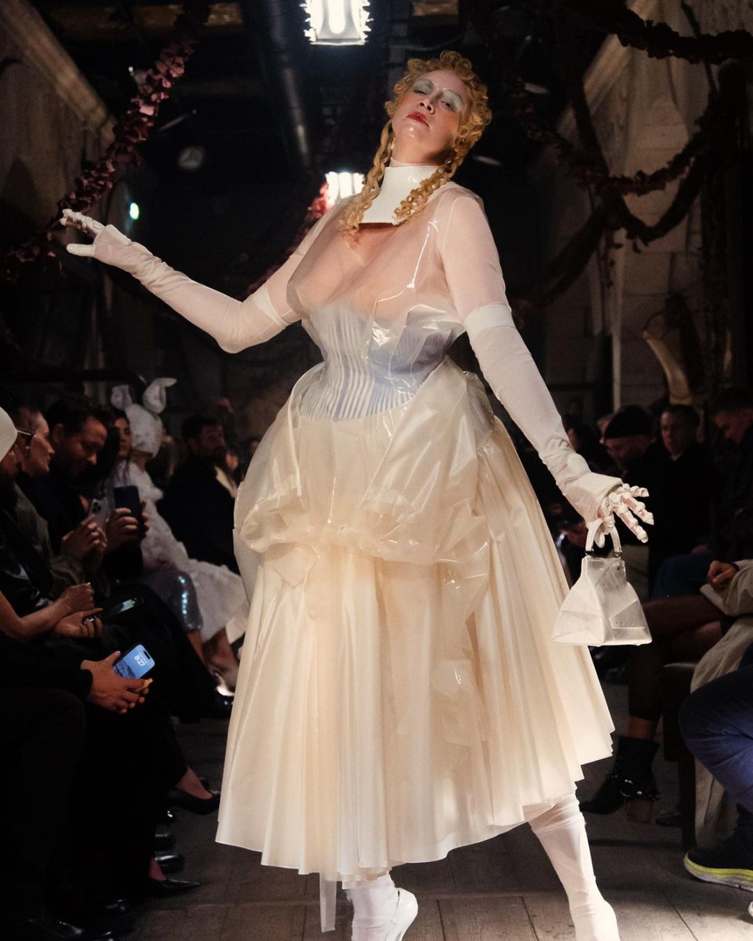 A woman in a translucent, avant-garde dress walks a runway during a fashion show, surrounded by an audience in a dimly lit space.