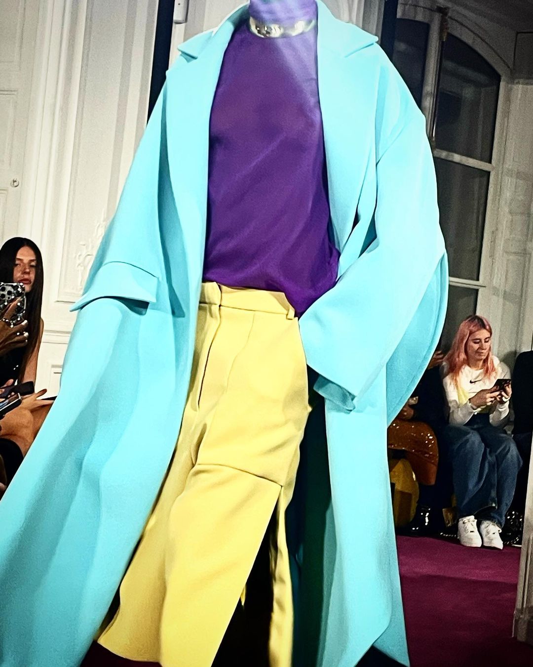 Fashion show presentation with a mannequin wearing a purple top, yellow skirt, and turquoise coat, in front of seated spectators.