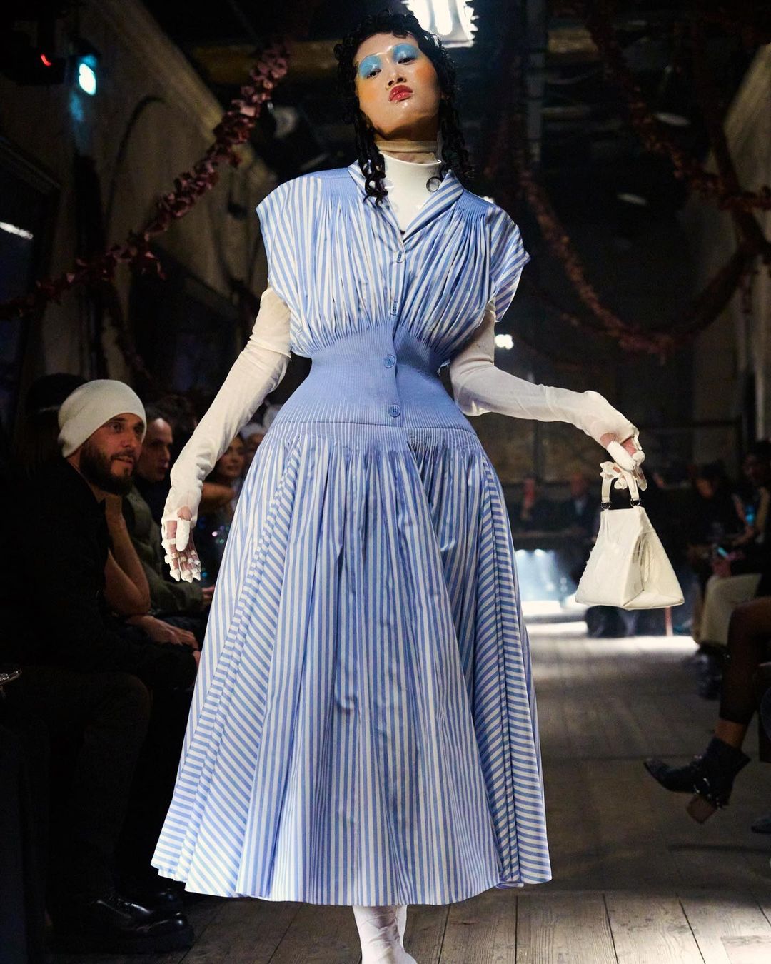 Fashion show runway featuring an adult female model in a blue and white striped dress, holding a white purse, walking toward the camera.