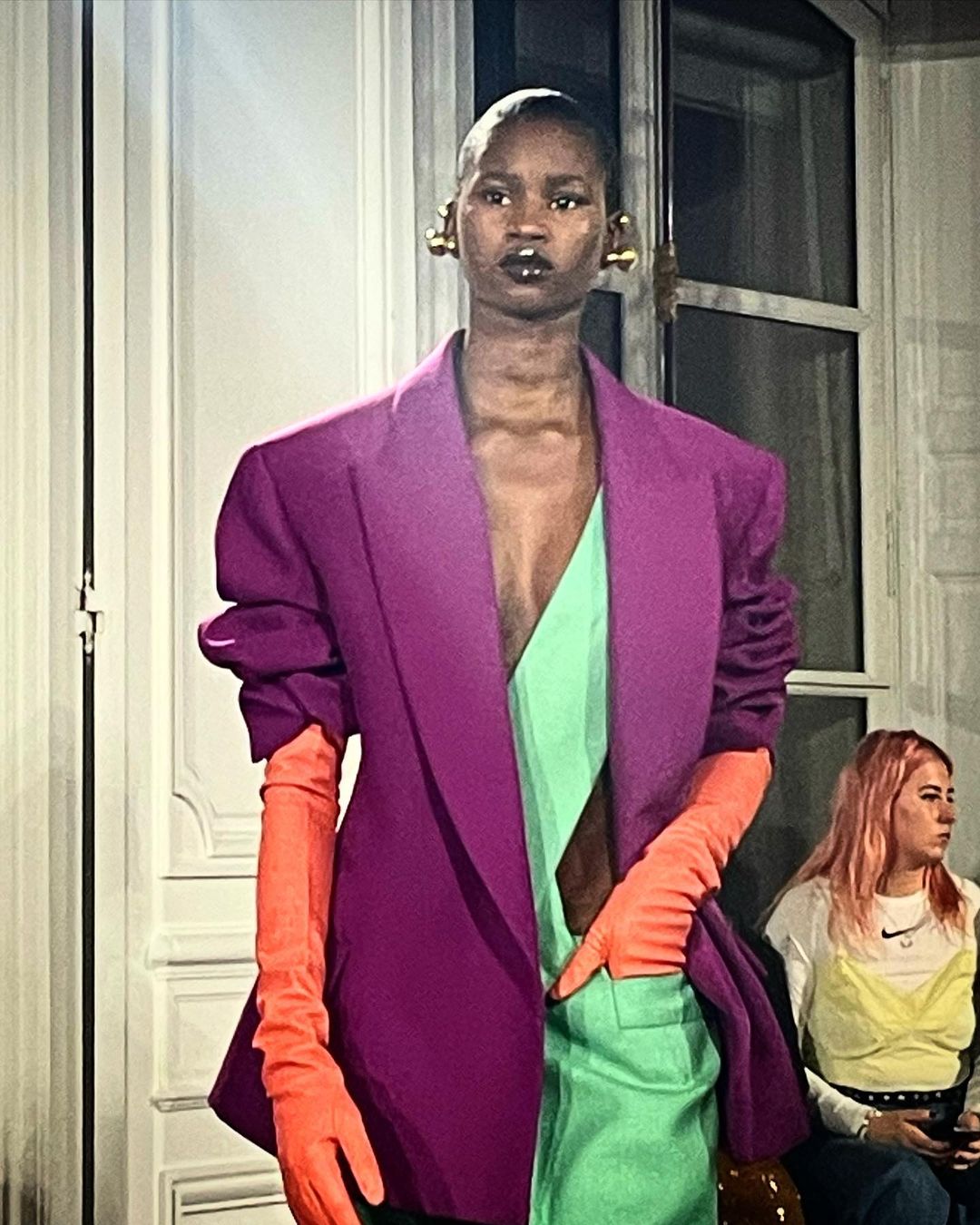 Runway fashion show featuring a Black model wearing a magenta blazer, chartreuse top and pants, and bright orange gloves.