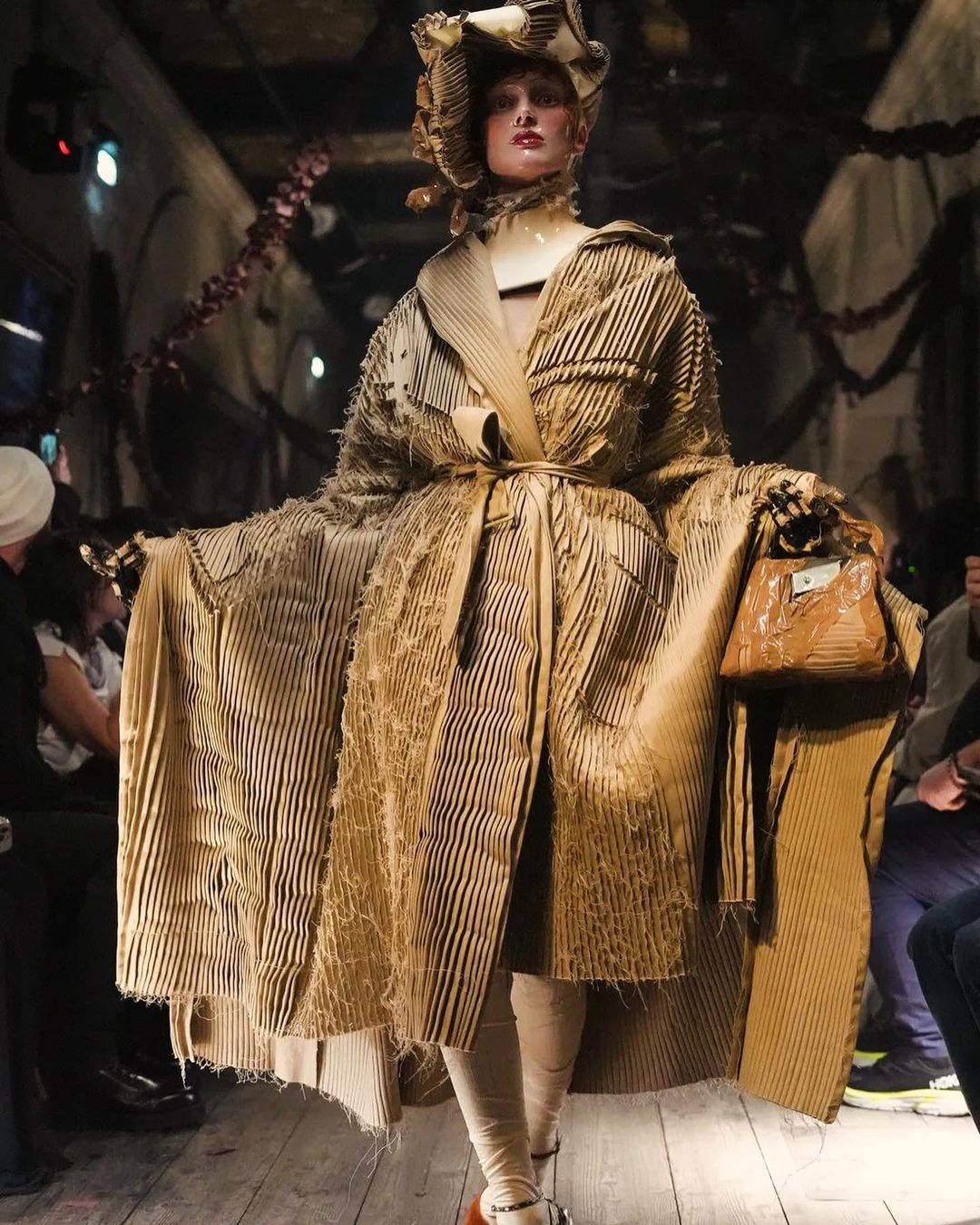 Runway fashion model in a sculptural coat, hat, and handbag made from cardboard. A tan corrugated cardboard outfit in a fashion show.