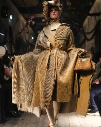 Model wearing a structured pleated brown coat and matching hat holding a handbag on a runway