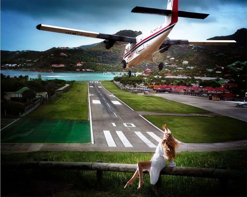 Small airplane flying low over a runway next to green hills and ocean as a woman sits on a rail in the foreground.