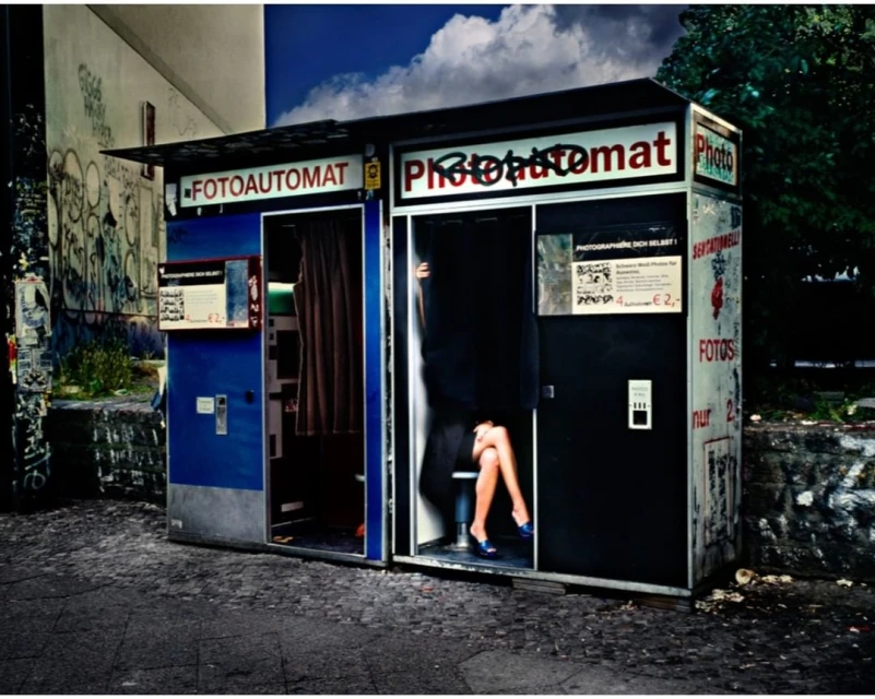 Photo booth in an urban outdoor setting featuring a woman's legs and blue shoes visible within one of the photo booth compartments.