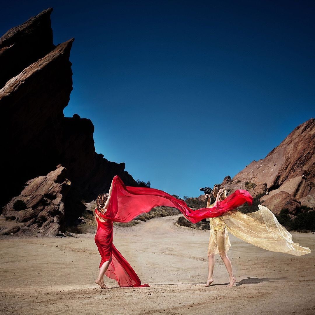 Two women in draped fabric pose in the desert with rocky formations under a clear blue sky, capturing a blend of fashion and nature.
