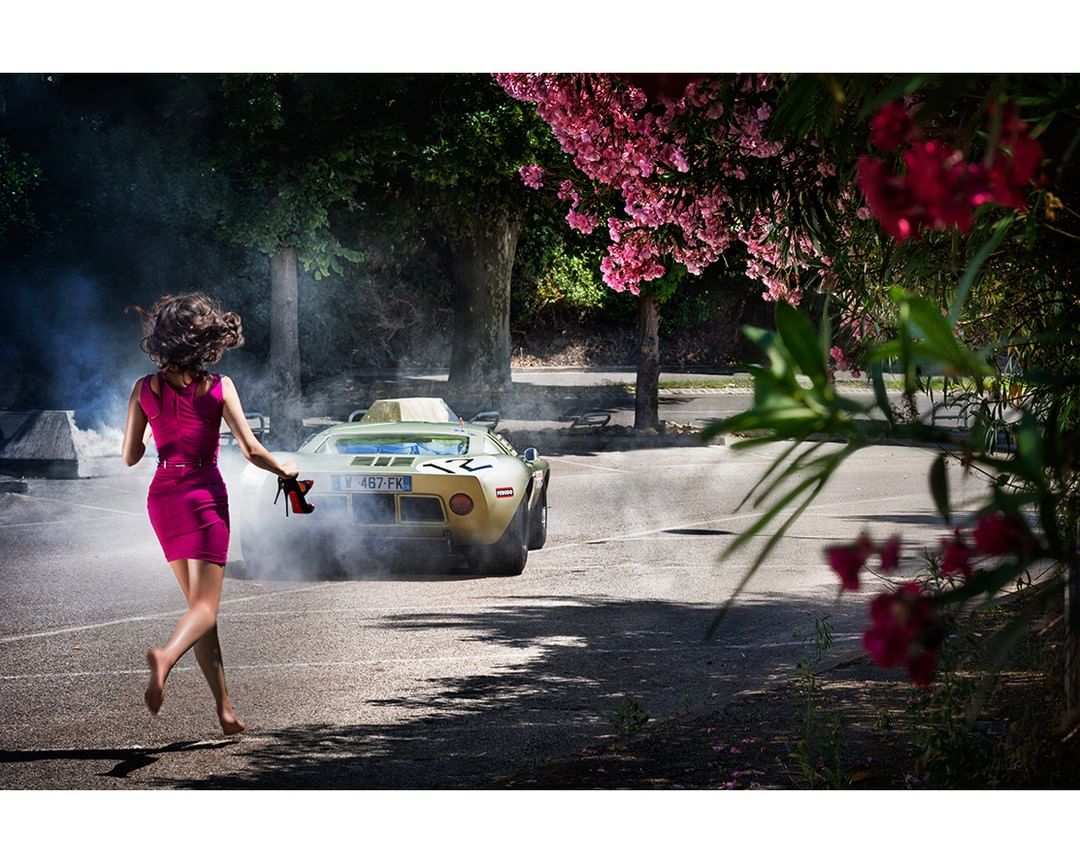 A barefoot woman in a pink dress runs on asphalt, clutching red high heels, away from a smoking beige sports car amidst lush greenery.