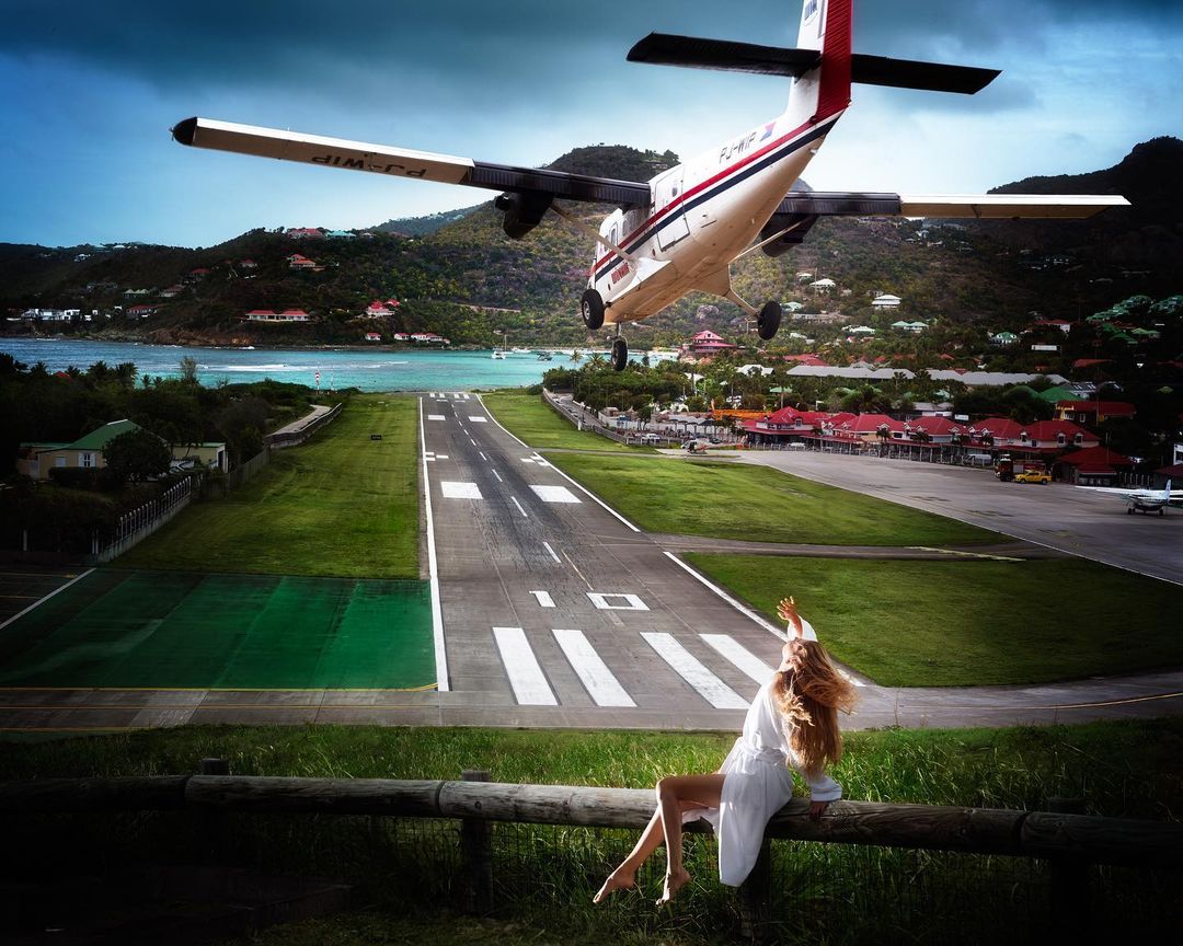 Airplane landing over tropical island runway as a woman sits on a log in the foreground watching the incoming plane. Sunny sky day.