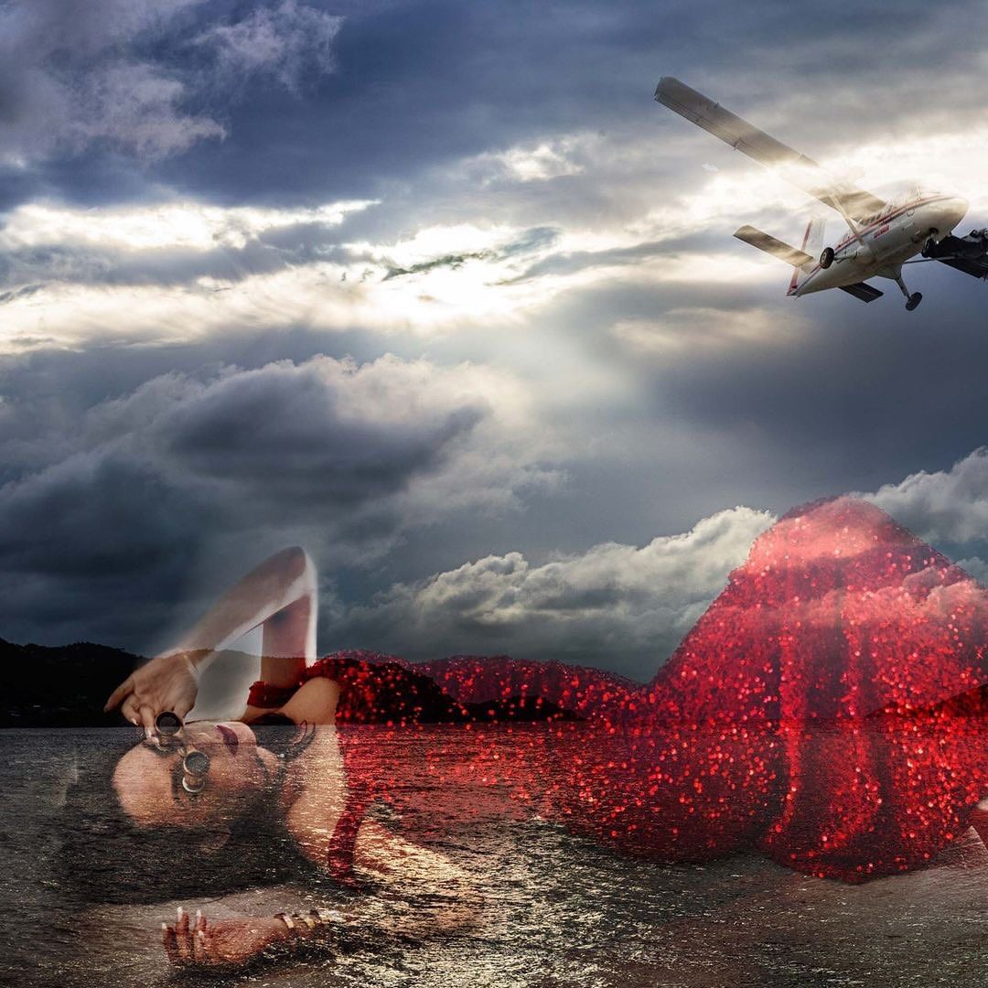 Woman in red dress reclines at water's edge with dramatic sky and airplane overhead, creating a surreal, fashion-forward visual.