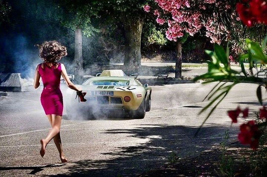 A woman in a magenta dress runs barefoot on asphalt towards a classic car in a smoky outdoor setting with pink blossoms.