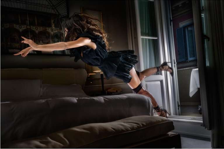 Woman in black dress seemingly jumps from bed to balcony in dark, atmospheric bedroom. Interior with contrasting lighting.