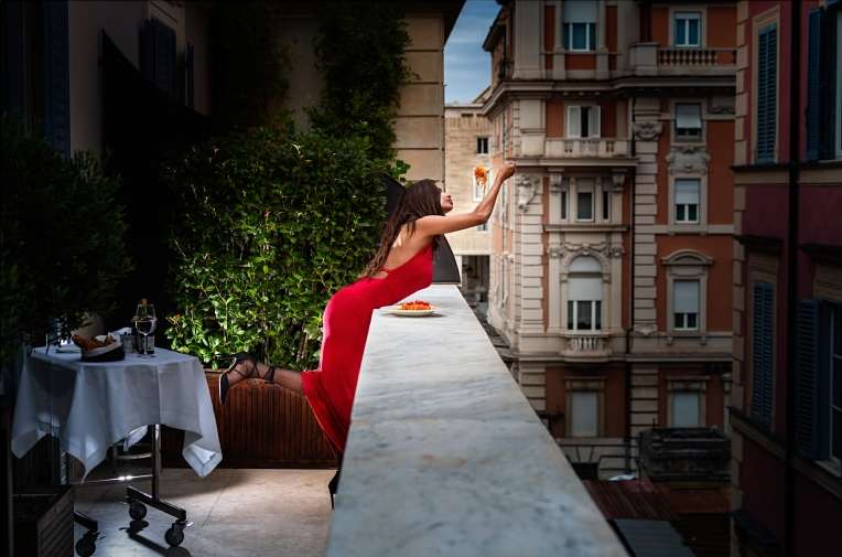 Woman in red dress reaches from balcony towards buildings with pasta in hand. Food, buildings, balcony, greenery, outdoor scene.
