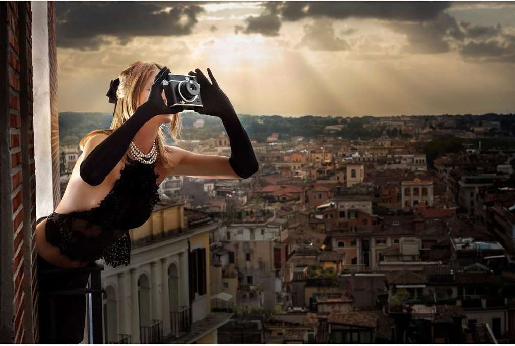 Woman with camera stands on balcony overlooking city at sunset. She wears black gloves, pearls, and black lace. Urban scene with warm light.