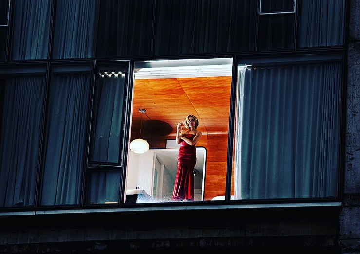 Elegant woman in a red gown stands inside a modern apartment, framed by a large window with dark blue curtains at nighttime.
